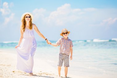 Happy beautiful mother in long white dress enjoying beach time with her son wearing hat and sunglasses