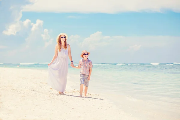 Happy beautiful mother in long white dress enjoying beach time with her son wearing hat and sunglasses — Zdjęcie stockowe