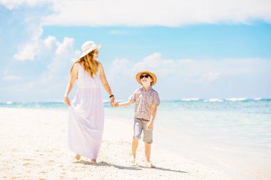 Happy beautiful mother in long white dress enjoying beach time with her son wearing hat and sunglasses