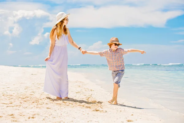 Happy beautiful mother in long white dress enjoying beach time with her son wearing hat and sunglasses — Stok fotoğraf