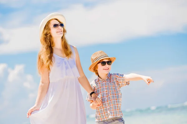 Mãe bonita feliz em vestido branco longo desfrutando de tempo de praia com seu filho vestindo chapéu e óculos de sol — Fotografia de Stock