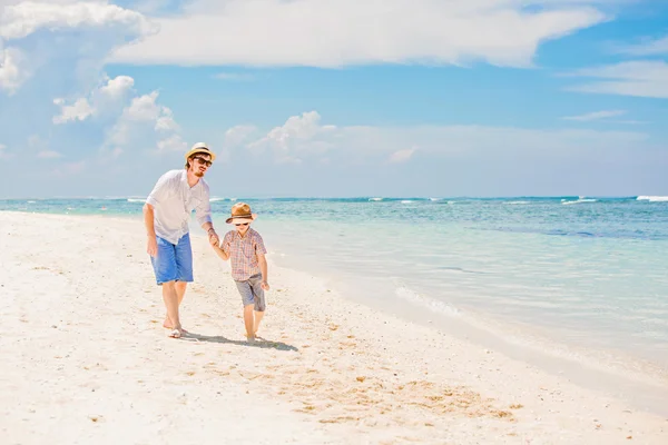 Young happy father and his little son enjoing summer time walking barefoot at the beach with ocean, beautiful clouds on background — Zdjęcie stockowe