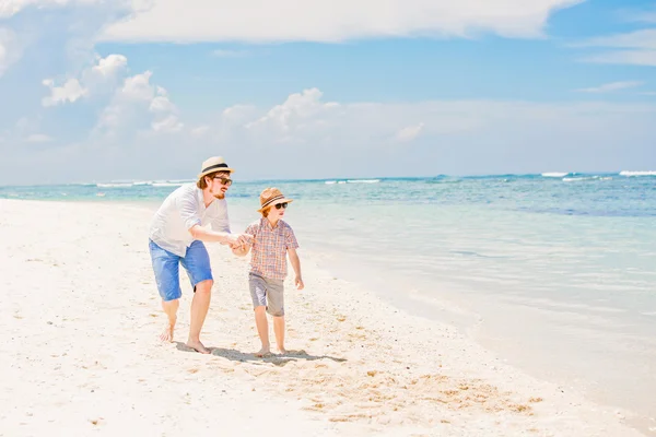 Young happy father and his little son enjoing summer time walking barefoot at the beach with ocean, beautiful clouds on background — Stok fotoğraf