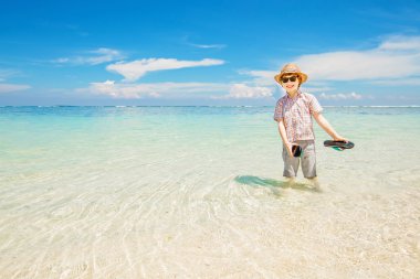 Happy kid boy wearing summer hat and hipster sunglasses walks in shallow water enjoying sunny day