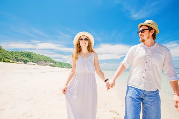 Jovem casal feliz em chapéus e óculos de sol caminhar na praia tropical com areia branca contra bela vista sobre o fundo — Fotografia de Stock