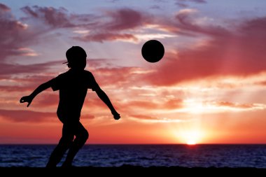 Silhouette of a boy playing football or soccer at the beach with beautiful sunset background