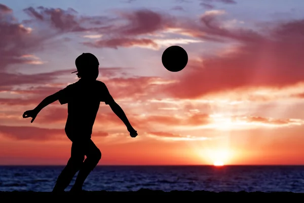 Silhouette of a boy playing football or soccer at the beach with beautiful sunset background