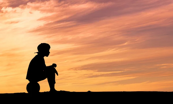 Silhouette of a boy sitting on football or soccer ball at the beach with sunset background