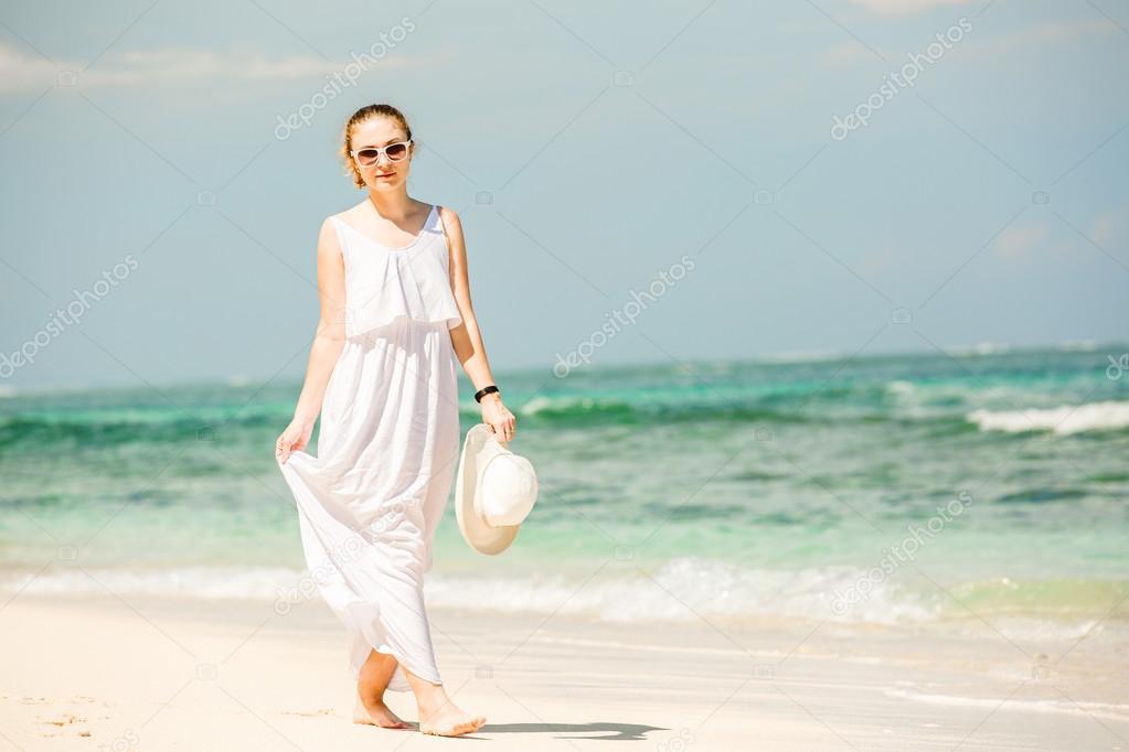 Young woman in long white dress holding hat walks along tropical beach having great summer time on holidays
