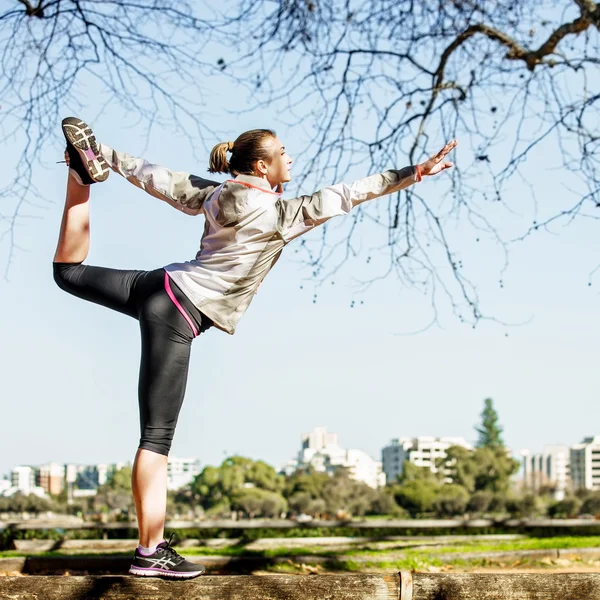 Jeune femme s'étirant sur le banc avant de faire du jogging dans la nature d'automne avec la ville en arrière-plan — Photo