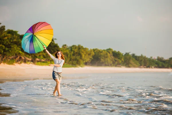 Jovencita alegre con paraguas de arco iris divirtiéndose en la playa antes del atardecer. Viajes, vacaciones, vacaciones, concepto de estilo de vida saludable — Foto de Stock