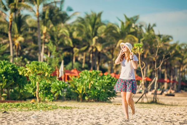 Beautiful young caucasian woman at the beach in white hat talking on mobile phone and laughing — Stockfoto