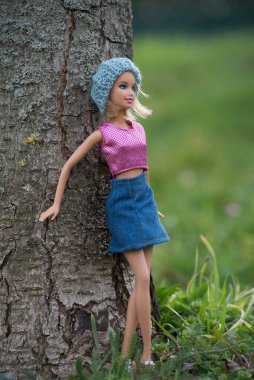 Mulhouse - France - 4 march 2021 - Portrait of blond Barbie doll with woolen hat and blue jeans skirt standing in a public garden against a tree trunk