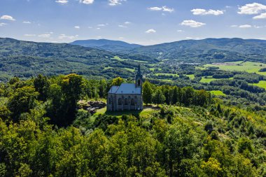 Çek Cumhuriyeti 'nin Bozi Hora tepesindeki tarihi kilisesinin, geleneksel çatılarının, yeşilliğin çevresindeki Zulova kasabasının insansız hava aracı görüntüsü ve gün ışığında yakalanan huzurlu kasaba düzeni..