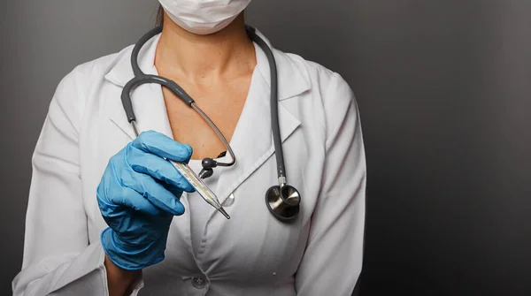 Tıbbi maske takmış bir kadın doktor ve beyaz laboratuvar önlüğü elinde bir termometre tutuyor. Virüsü, Covid19 'u, salgını, koronavirüsü, sağlık hizmetlerini ve doktorların konseptini engelleyin. İzole edilmiş. Boşluğu kopyala.