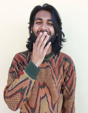 Yakışıklı bir gencin portresi, gülüyor ve elleriyle ağzını kapatıyor, sakallı ve uzun saçlı Hintli genç bir adamın stüdyo fotoğrafı.