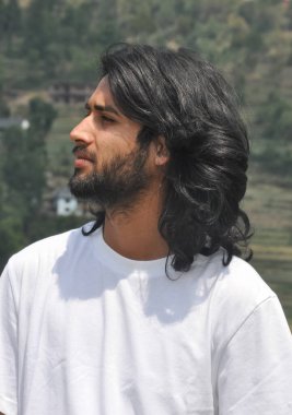 Yakışıklı, sakallı, uzun saçlı, yana bakan genç bir adamın, dışarıda beyaz t-shirt giyen genç bir adamın yakın plan fotoğrafı.