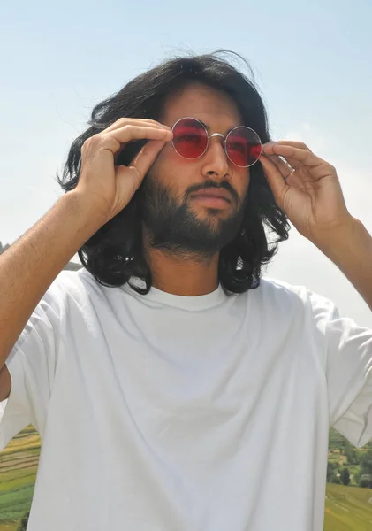 A long haired young men wearing sunglass with his hands with looking sideways, A good looking bearded guy standing outside with wearing white t-shirt