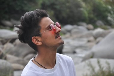 Nehrin kenarında beyaz tişört ve kırmızı güneş gözlüğü takmış yukarı bakan Hintli genç bir adam.