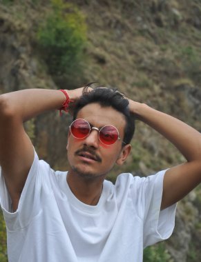 Beyaz tişörtlü, kırmızı gözlüklü, dışarıda poz veren, ellerini yana doğru uzatan seksi bir gencin portresi.