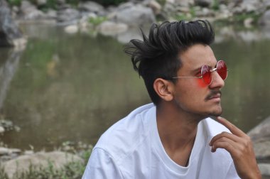 Kuzey Hintli genç bir adam nehir kenarında oturuyor, çenesinde parmak, yana bakıyor, beyaz tişörtlü ve kırmızı gözlüklü bir adam. 