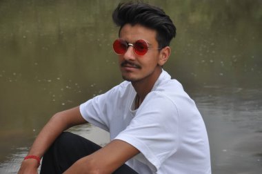 Nehrin kenarında oturan yakışıklı genç bir adam elini yan yatmış dizine yaslamış beyaz tişörtlü ve kırmızı gözlüklü Hintli bir adam. 