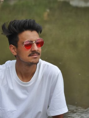 Nehrin kenarında oturan düşünceli genç bir adamın yakın plan fotoğrafı beyaz tişörtlü ve kırmızı gözlüklü Hintli bir adam. 
