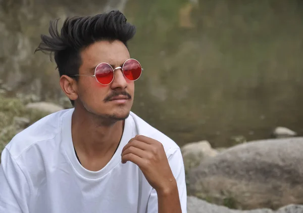 北インドのハンサムな若い男が横に見て川の横に座って 白いTシャツと赤いサングラスを身に着けている男 — ストック写真