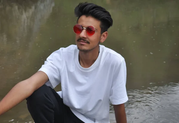 一个英俊的年轻人坐在河边 手放在膝盖上 侧面看去 一个穿着白色T恤和红色太阳镜的印度人 — 图库照片