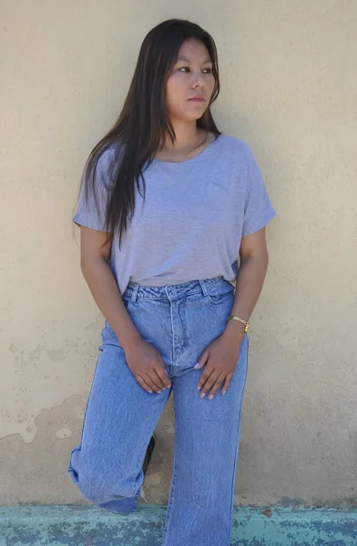 A south asian young girl posing with wearing gray tshirt and blue jean standing against yellow wall with looking sideways