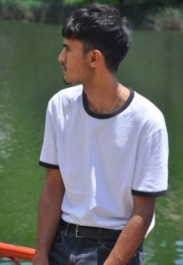 Beyaz tişört ve siyah kot giyen yakışıklı Hintli genç göl kenarındaki güvenlik bariyerinde oturuyor ve yan bakıyor. 