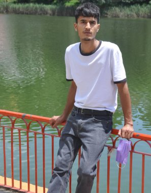 Beyaz tişört ve siyah kot pantolon giyen, elinde maskeyle gölün kenarındaki güvenlik bariyerine oturmuş kameraya bakan yakışıklı bir Kuzey Hintli genç. 