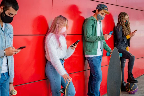 Young people wearing face safety masks using smart mobile phones while keeping social distance during coronavirus time - Technology and covid-19 spread prevention concept