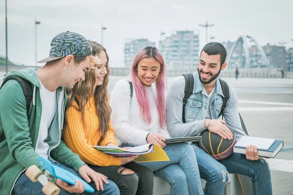 多民族学生冗談と一緒に大学で屋外のベンチに座って話 本やタブレットで勉強幸せな若い十代の若者のグループ — ストック写真