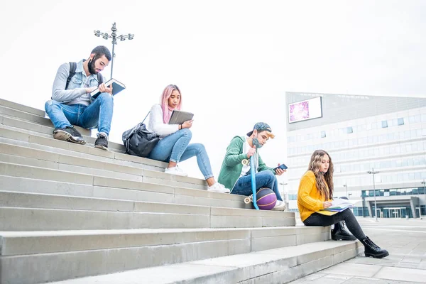 コロナウイルスのパンデミックの間に屋外で勉強する若者の大学 社会的距離を保つ階段に屋外に座って保護マスクを持つ4人の多国籍大学の学生 — ストック写真