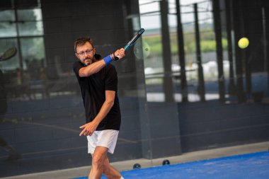 Kapalı alanda padel oynayan genç sporcu çocuk raketle topa vuruyor.