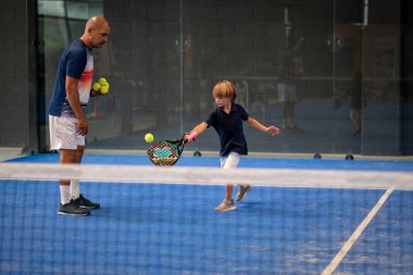 Öğrencisi Padel 'den çocuğa ders verirken gözlemci, küçük çocuğa kapalı tenis kortunda nasıl padel oynanacağını öğretiyor.
