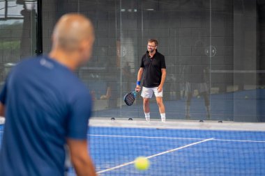 Öğrencisi Padel 'in erkek öğrencisine ders verirken gözlemci çocuğa kapalı tenis kortunda nasıl Padel oynanacağını öğretiyor.
