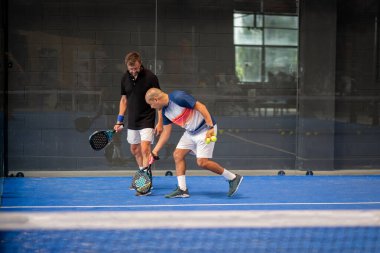 Öğrencisi Padel 'in erkek öğrencisine ders verirken gözlemci çocuğa kapalı tenis kortunda nasıl Padel oynanacağını öğretiyor.