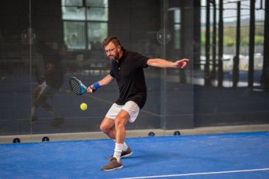 Kapalı alanda padel oynayan genç sporcu çocuk raketle topa vuruyor.