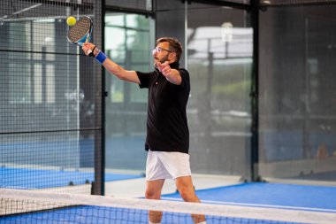 Kapalı alanda padel oynayan genç sporcu çocuk raketle topa vuruyor.