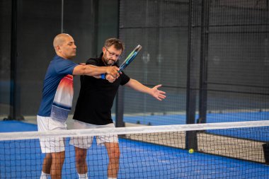 Öğrencisi Padel 'in erkek öğrencisine ders verirken gözlemci çocuğa kapalı tenis kortunda nasıl Padel oynanacağını öğretiyor.