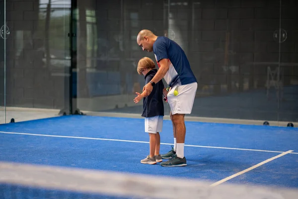 Öğrencisi Padel 'den çocuğa ders verirken gözlemci, küçük çocuğa kapalı tenis kortunda nasıl padel oynanacağını öğretiyor.