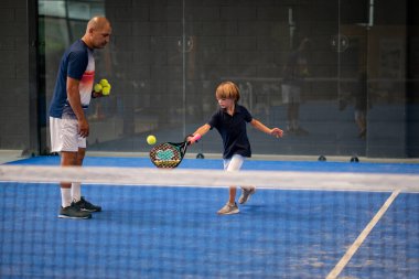 Öğrencisi Padel 'den çocuğa ders verirken gözlemci, küçük çocuğa kapalı tenis kortunda nasıl padel oynanacağını öğretiyor.