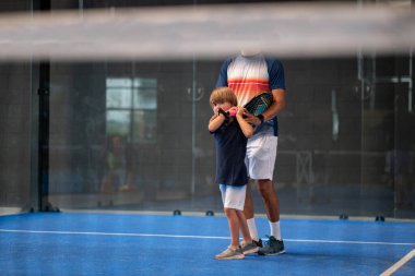 Öğrencisi Padel 'den çocuğa ders verirken gözlemci, küçük çocuğa kapalı tenis kortunda nasıl padel oynanacağını öğretiyor.