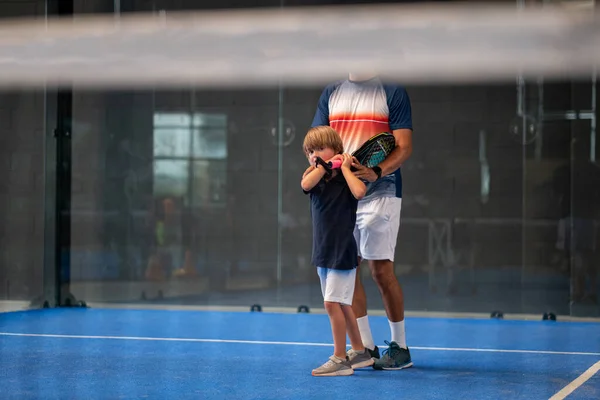 Öğrencisi Padel 'den çocuğa ders verirken gözlemci, küçük çocuğa kapalı tenis kortunda nasıl padel oynanacağını öğretiyor.