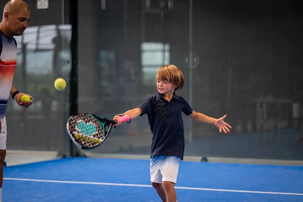 Öğrencisi Padel 'den çocuğa ders verirken gözlemci, küçük çocuğa kapalı tenis kortunda nasıl padel oynanacağını öğretiyor.