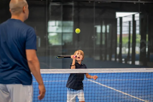 Öğrencisi Padel 'den çocuğa ders verirken gözlemci, küçük çocuğa kapalı tenis kortunda nasıl padel oynanacağını öğretiyor.