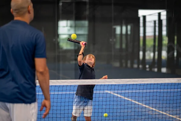 Öğrencisi Padel 'den çocuğa ders verirken gözlemci, küçük çocuğa kapalı tenis kortunda nasıl padel oynanacağını öğretiyor.