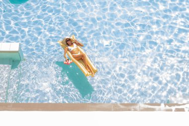 Havuzdaki şişme altın şiltenin üzerinde yüzen mayo ve şapkalı kız kokteyli. Yaz tatili ve rahatlama konsepti